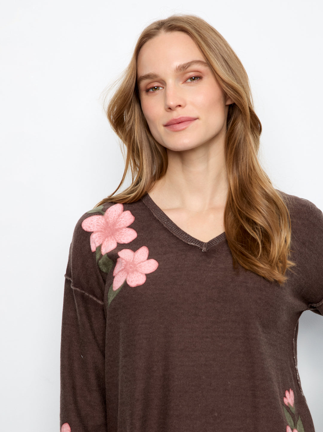 A woman wearing a dark brown long-sleeve V-neck top with pink floral embroidery on the shoulder, photographed against a plain white background.
