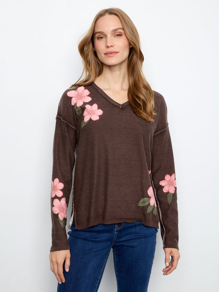 A woman wearing a brown long-sleeve V-neck knit top with pink floral embroidery on the shoulders and sleeves, paired with blue jeans, against a plain white studio background.