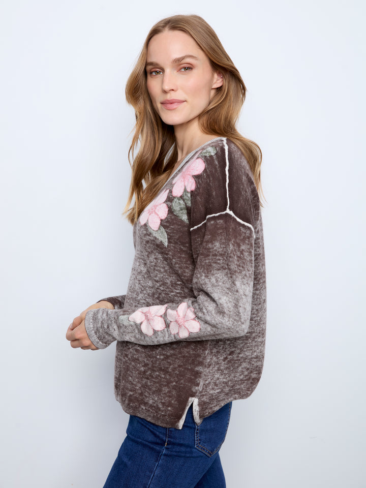 A woman modeling a brown and gray marled knit sweater with pink floral appliques on the chest and sleeve, paired with blue jeans against a light gray studio background.