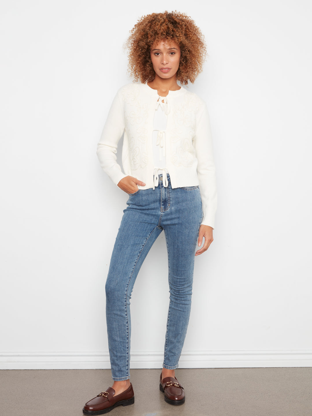 A woman stands against a white wall wearing a cream knit cardigan, blue skinny jeans, and brown leather loafers.