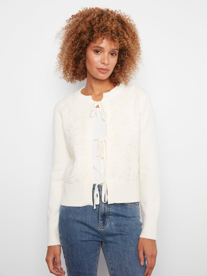 A woman with curly brown hair wearing a cream-colored knit cardigan with lace-up front details and blue jeans, standing against a plain white studio background.