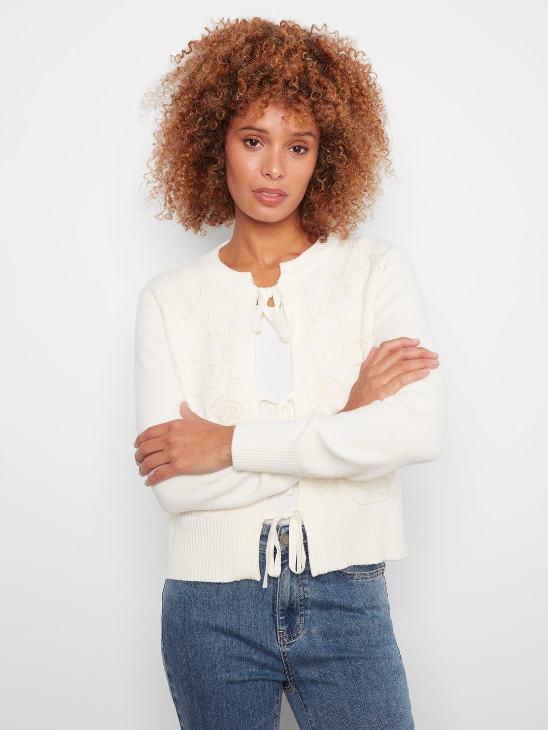 A woman with curly brown hair wearing a cream-colored knit cardigan with a tie-neck detail and blue jeans, standing against a plain white studio background.