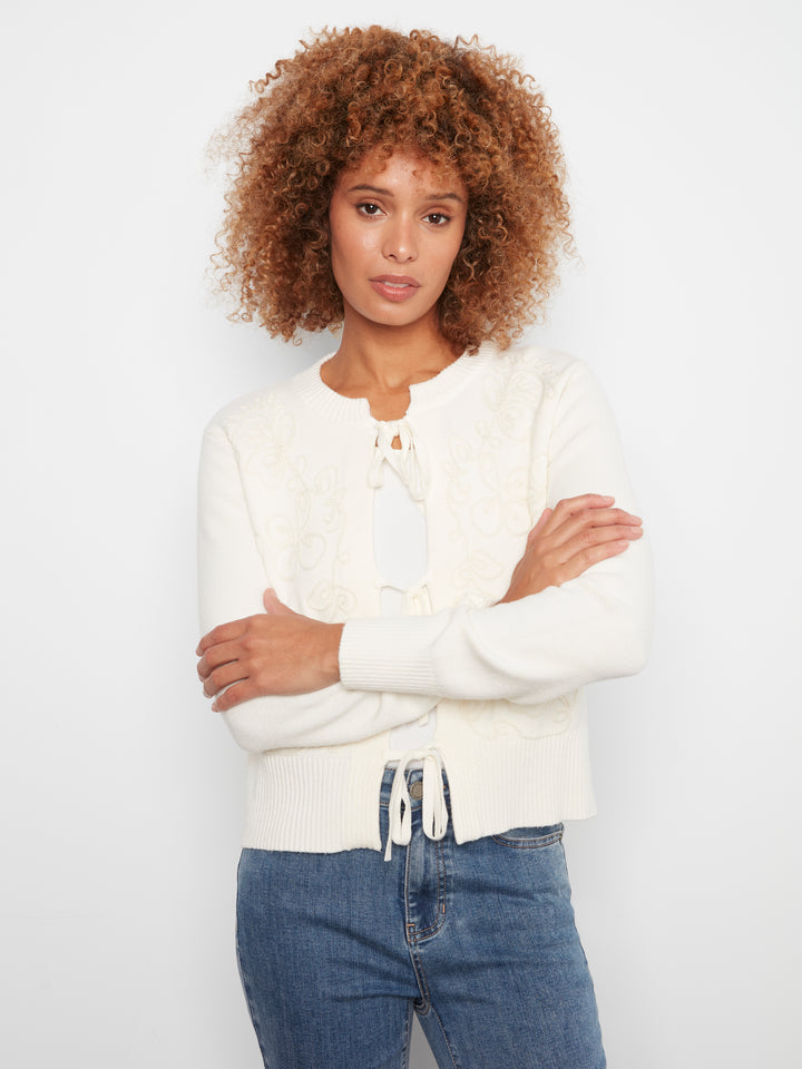 A woman with curly brown hair wearing a cream-colored knit cardigan with a tie-neck detail and blue jeans, standing against a plain white studio background.