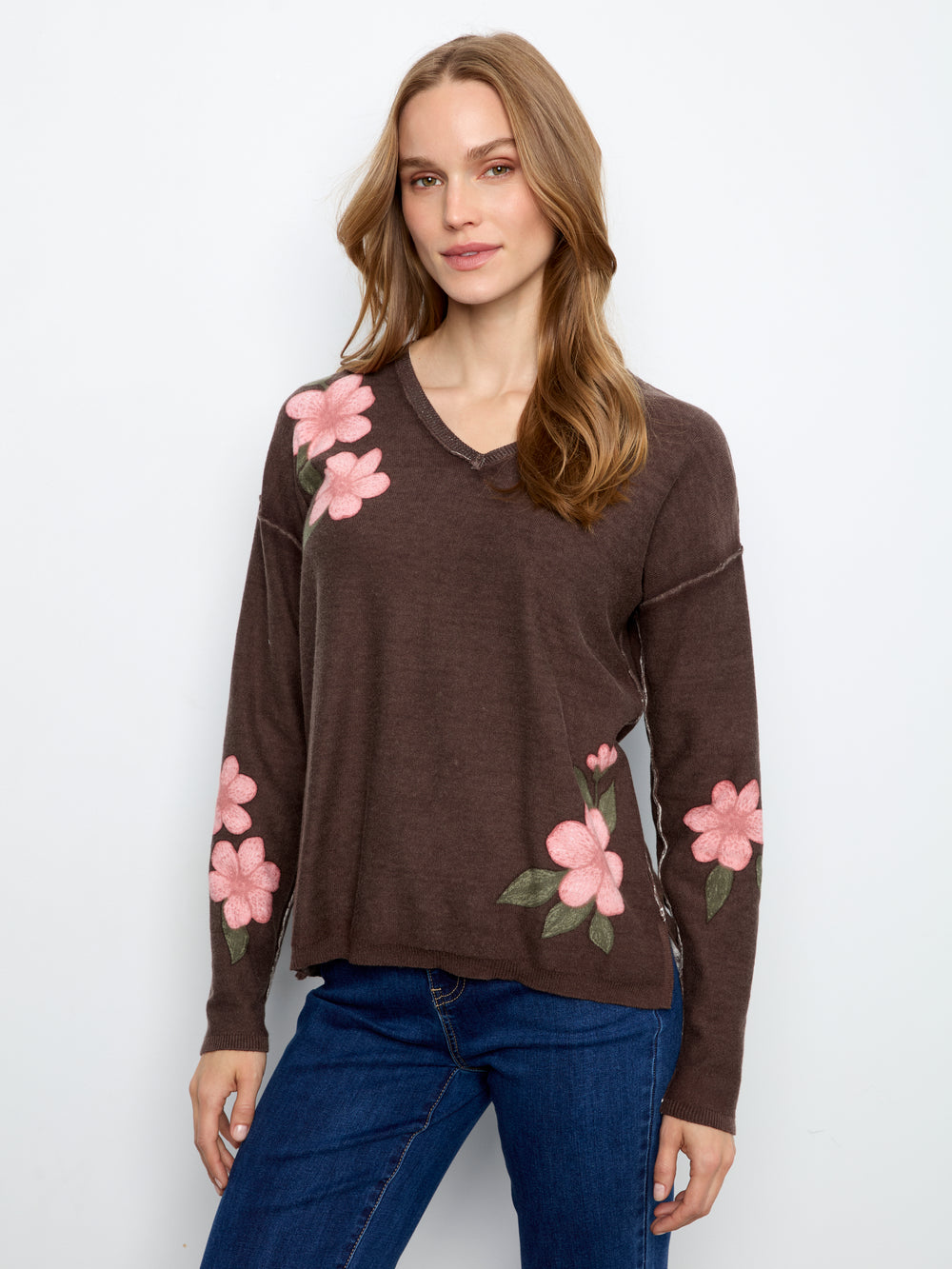 A woman wearing a brown long-sleeve V-neck sweater with pink floral embroidery on the shoulders and sleeves, paired with blue jeans, against a plain white background.