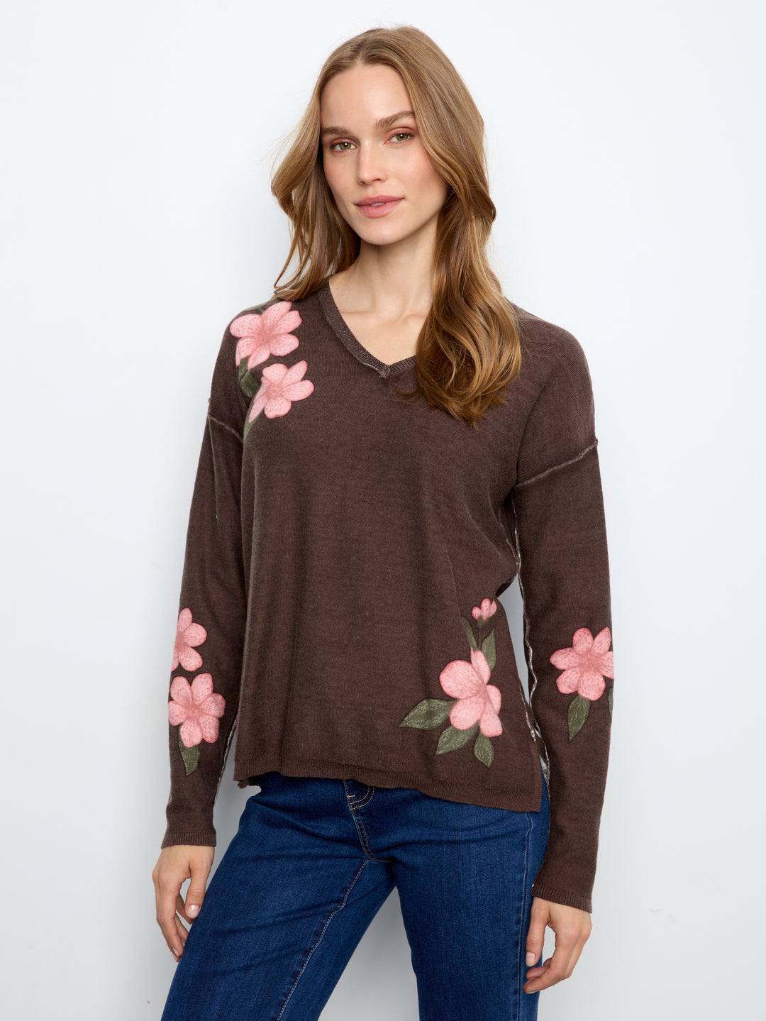 A woman wearing a brown long-sleeve V-neck sweater with pink floral embroidery on the shoulders and sleeves, paired with blue jeans, against a plain white background.