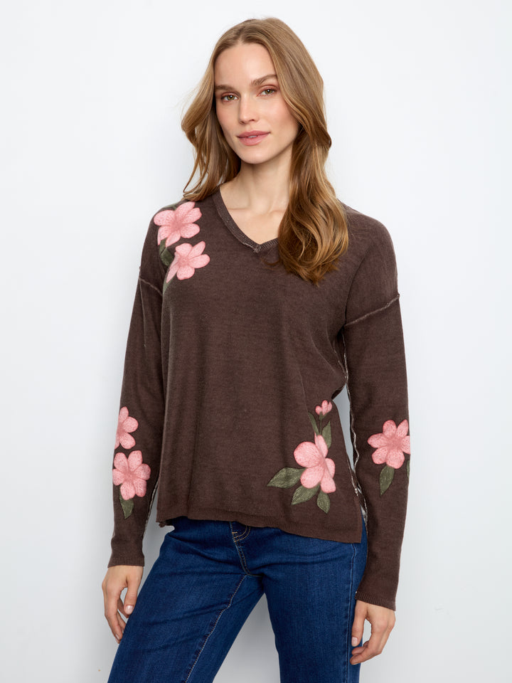 A woman wearing a brown long-sleeve V-neck sweater with pink floral embroidery on the shoulders and sleeves, paired with blue jeans, against a plain white background.