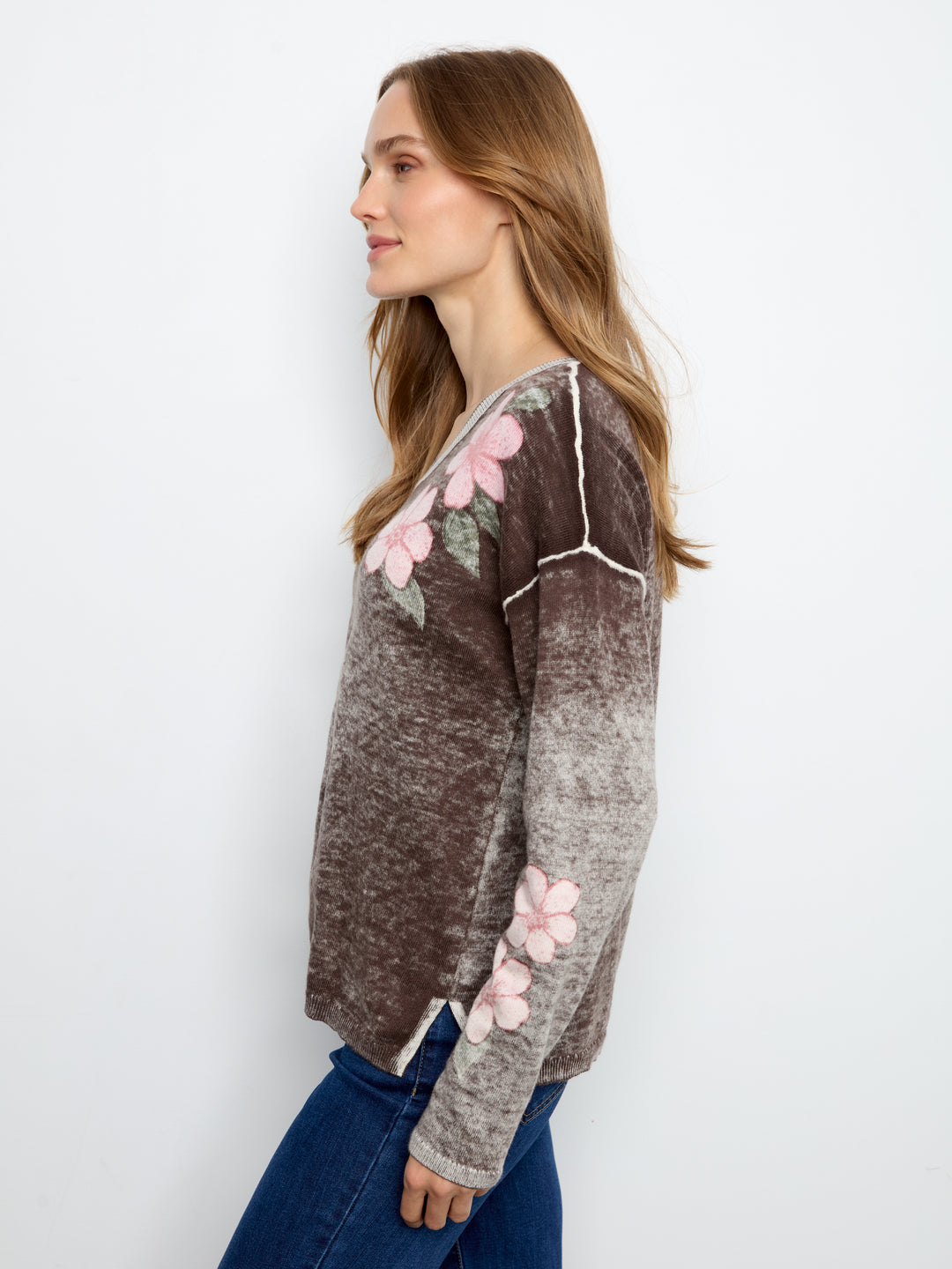 A woman in side profile wearing a brown and gray marled knit sweater with pink floral embroidery and white piping, paired with blue jeans, against a plain white background.