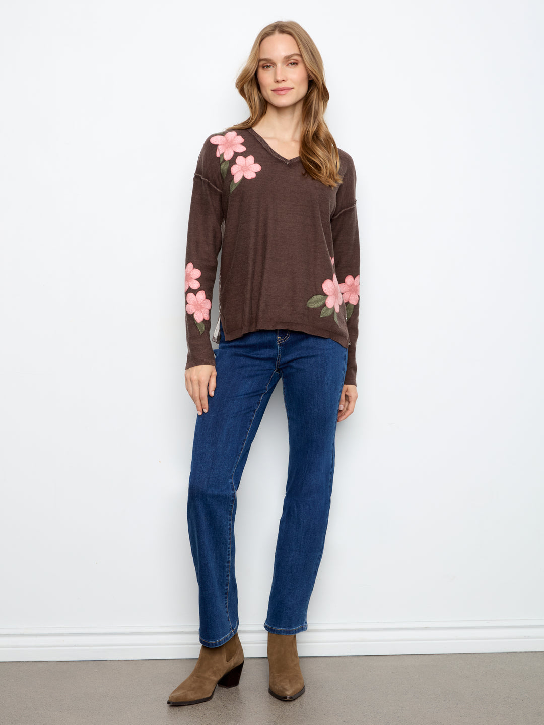 A woman modeling a brown long-sleeve V-neck sweater with pink floral embroidery on the shoulders and sleeves, paired with blue jeans and brown ankle boots, standing against a white wall.