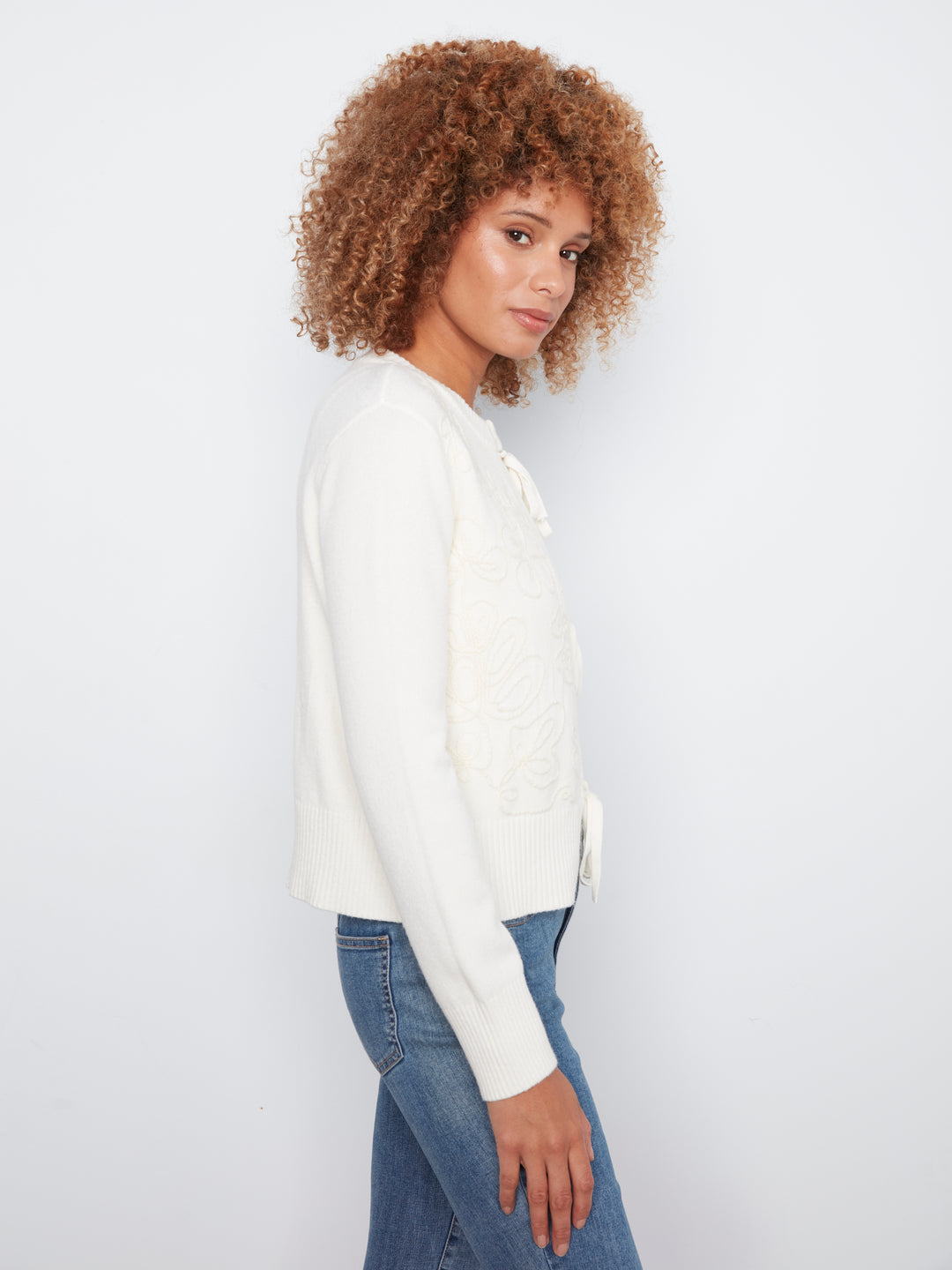A woman with curly brown hair is shown in profile wearing a white knit cardigan with textured details and blue jeans, set against a plain white studio background.