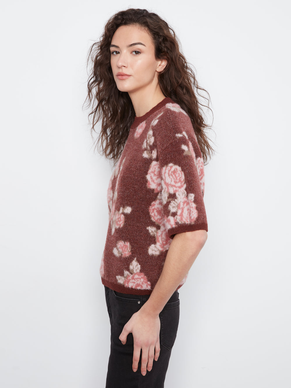 A woman wearing a short-sleeve crewneck knit sweater with a dusty rose and burgundy floral pattern, paired with black pants, photographed against a plain white studio background.