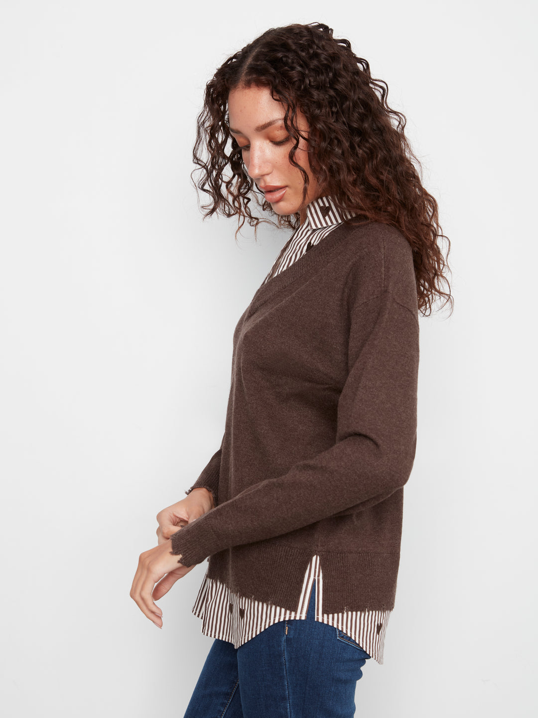 A woman with long, curly brown hair is shown in profile wearing a brown knit cardigan over a cream and black striped shirt, paired with blue jeans, against a plain light gray background.