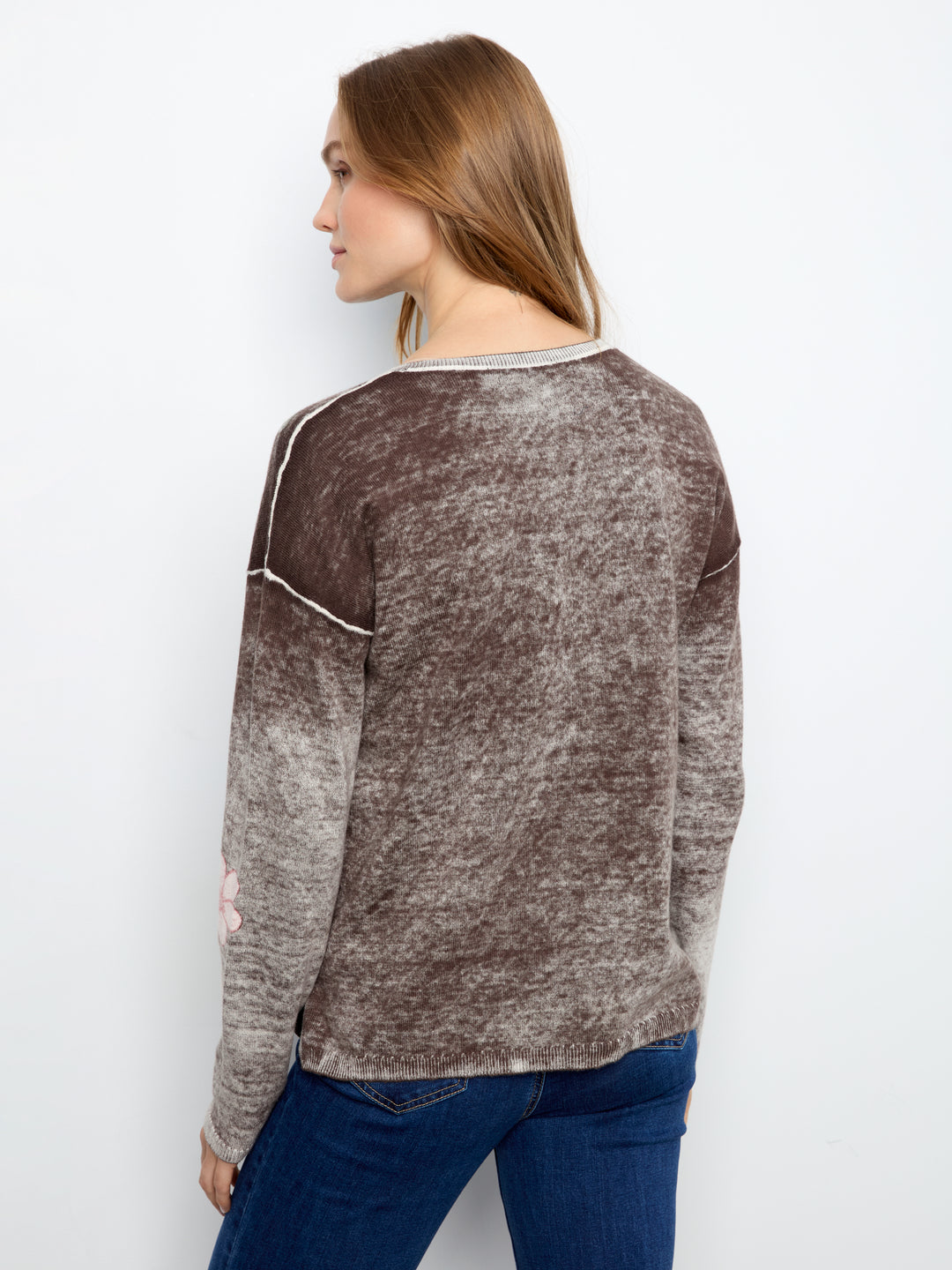 A woman shown from the back wearing a long-sleeve marled knit top in brown and gray tones with white contrast stitching, paired with blue jeans, against a plain white background.
