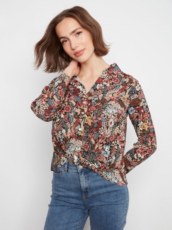 A woman wearing a long-sleeve button-up blouse with a dense multicolor floral print in red, blue, yellow, and beige tones, paired with blue jeans, against a plain light gray studio background.
