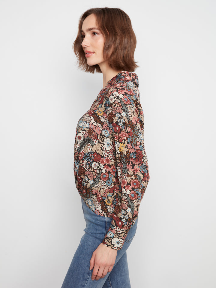 A woman with short brown hair is shown in profile wearing a long-sleeve crewneck top with a dense multicolor floral pattern, paired with blue jeans, against a plain light gray background.