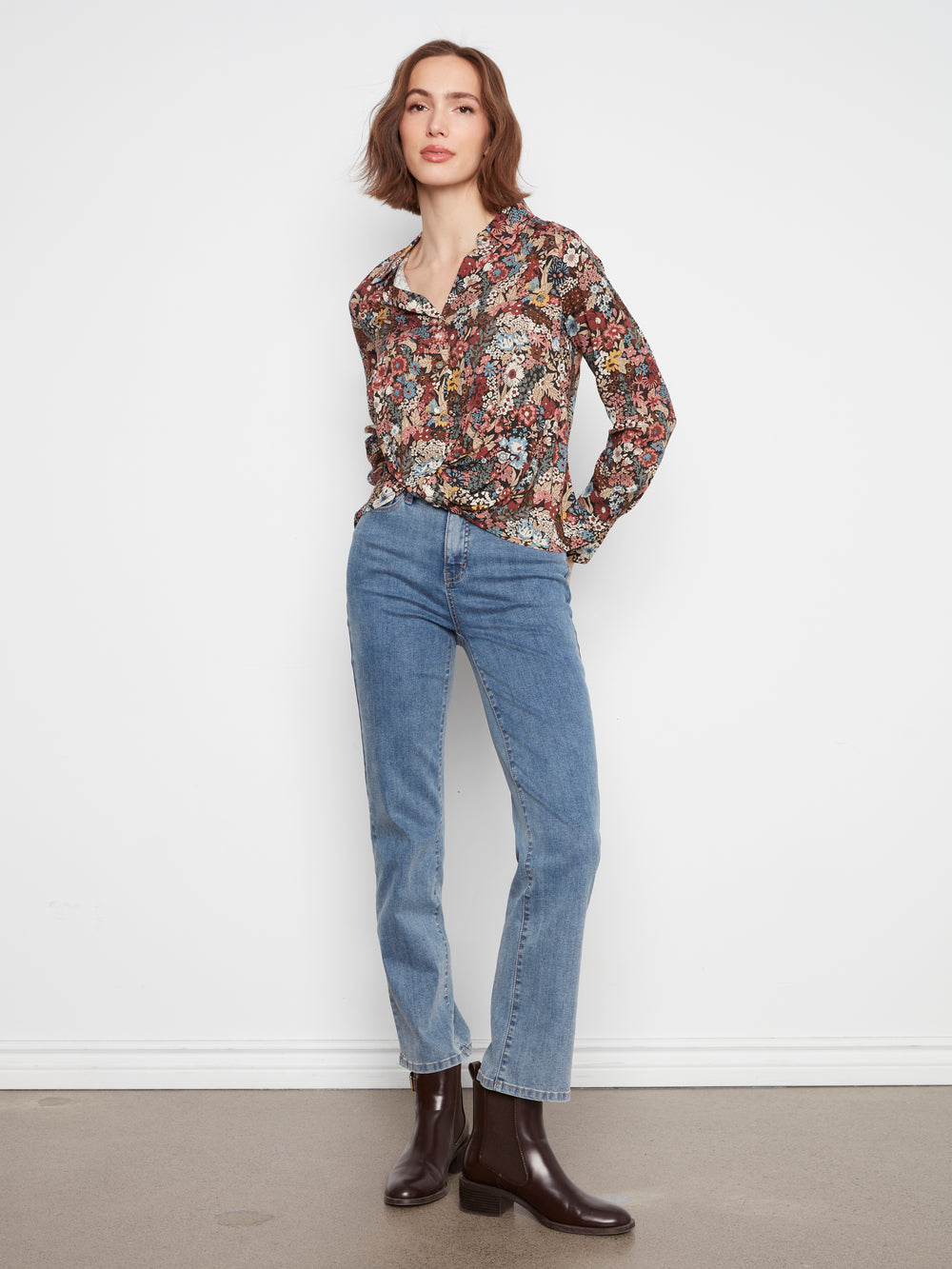 Woman wearing a floral blouse, blue jeans, and brown boots against a white background