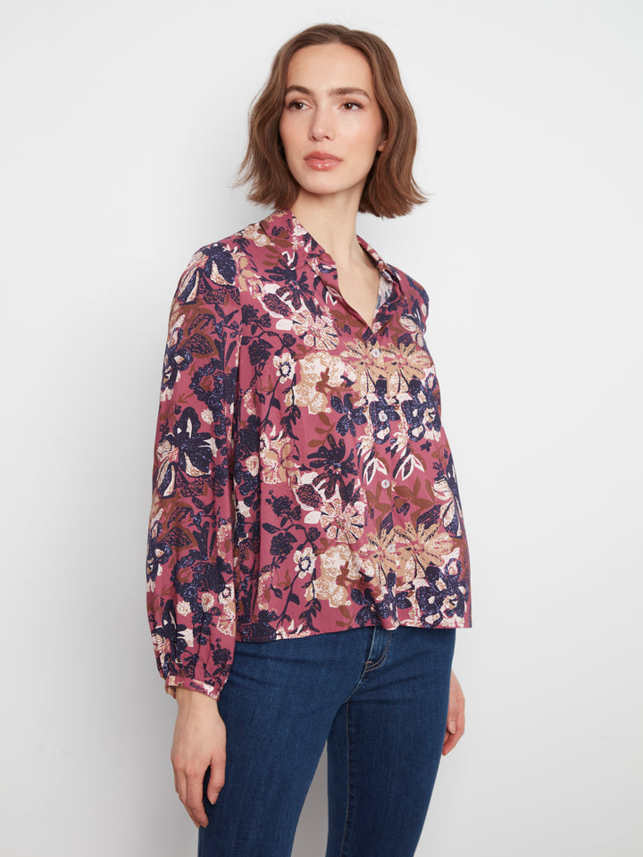 A woman wearing a long-sleeve button-up blouse with a dense floral and botanical print in dusty pink, burgundy, navy, and beige, paired with dark blue jeans, against a plain light gray studio background.