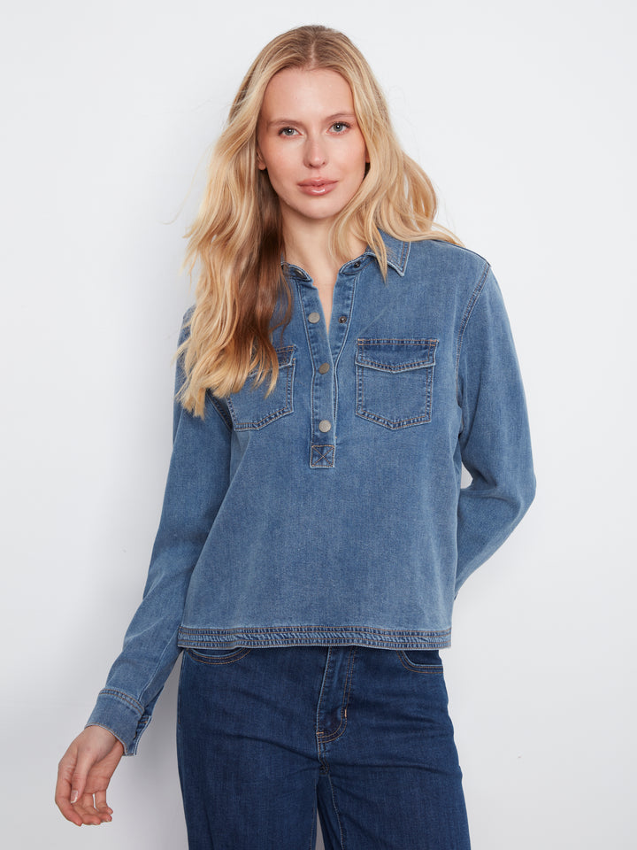 A woman modeling a medium-wash blue long-sleeve denim shirt with a buttoned henley-style placket and two chest pockets, paired with dark blue jeans against a plain light gray studio background.