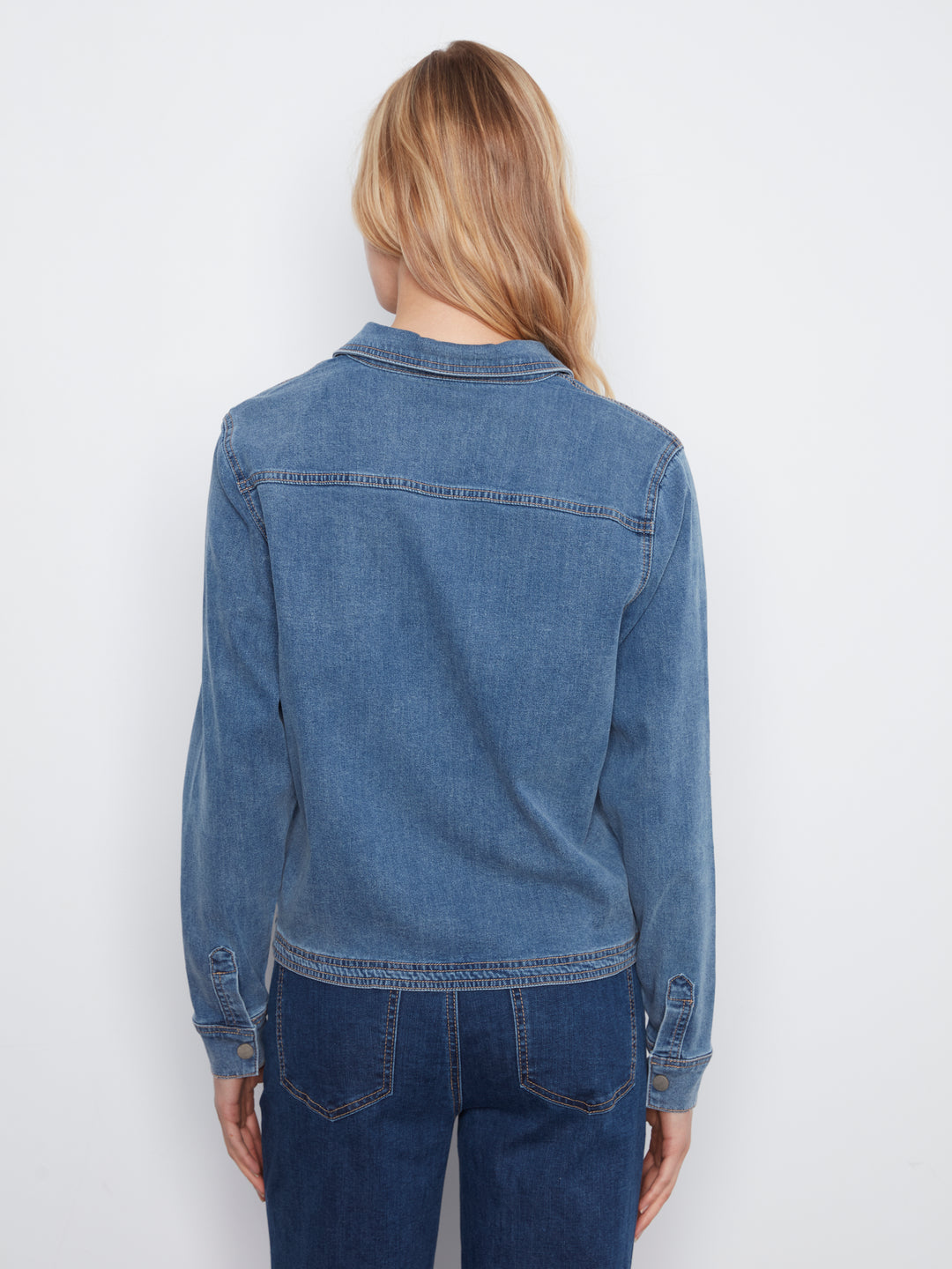 A woman viewed from the back wearing a medium-wash blue denim jacket and dark blue jeans against a plain white studio background.