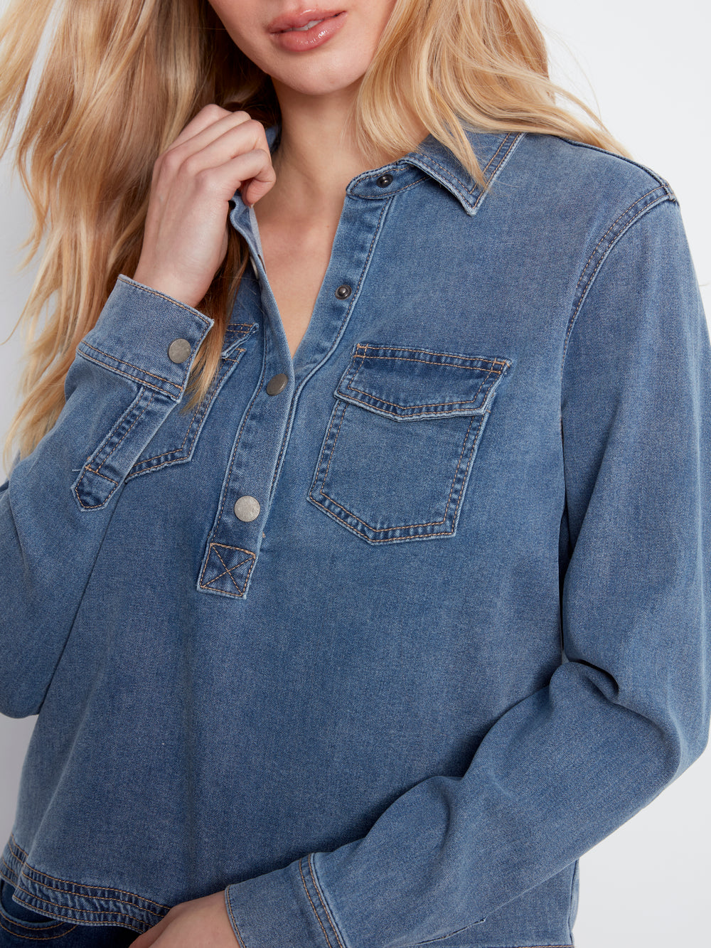 A close-up of a woman wearing a blue denim long-sleeve button-up shirt with chest pockets, shown against a plain light background.