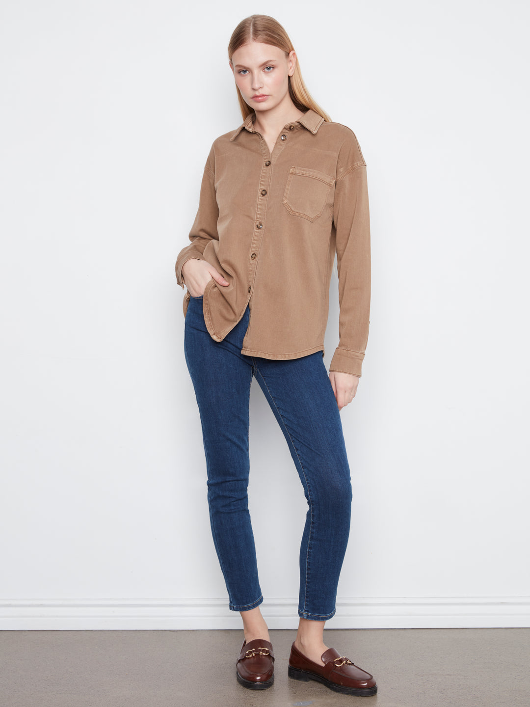A woman modeling a tan long-sleeve button-up shirt with chest pockets, paired with dark blue skinny jeans and brown loafers, standing against a white wall.