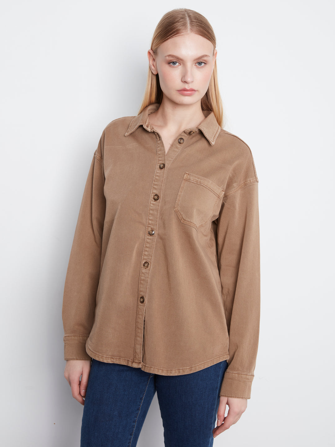 A woman modeling a long-sleeve button-up shirt in a warm tan or camel color, paired with dark blue jeans, against a plain light gray studio background.