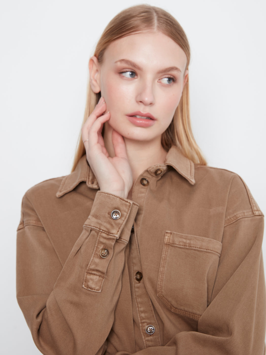 A blonde woman wearing a brown button-up shirt with a collar and chest pocket, posing against a light gray background.