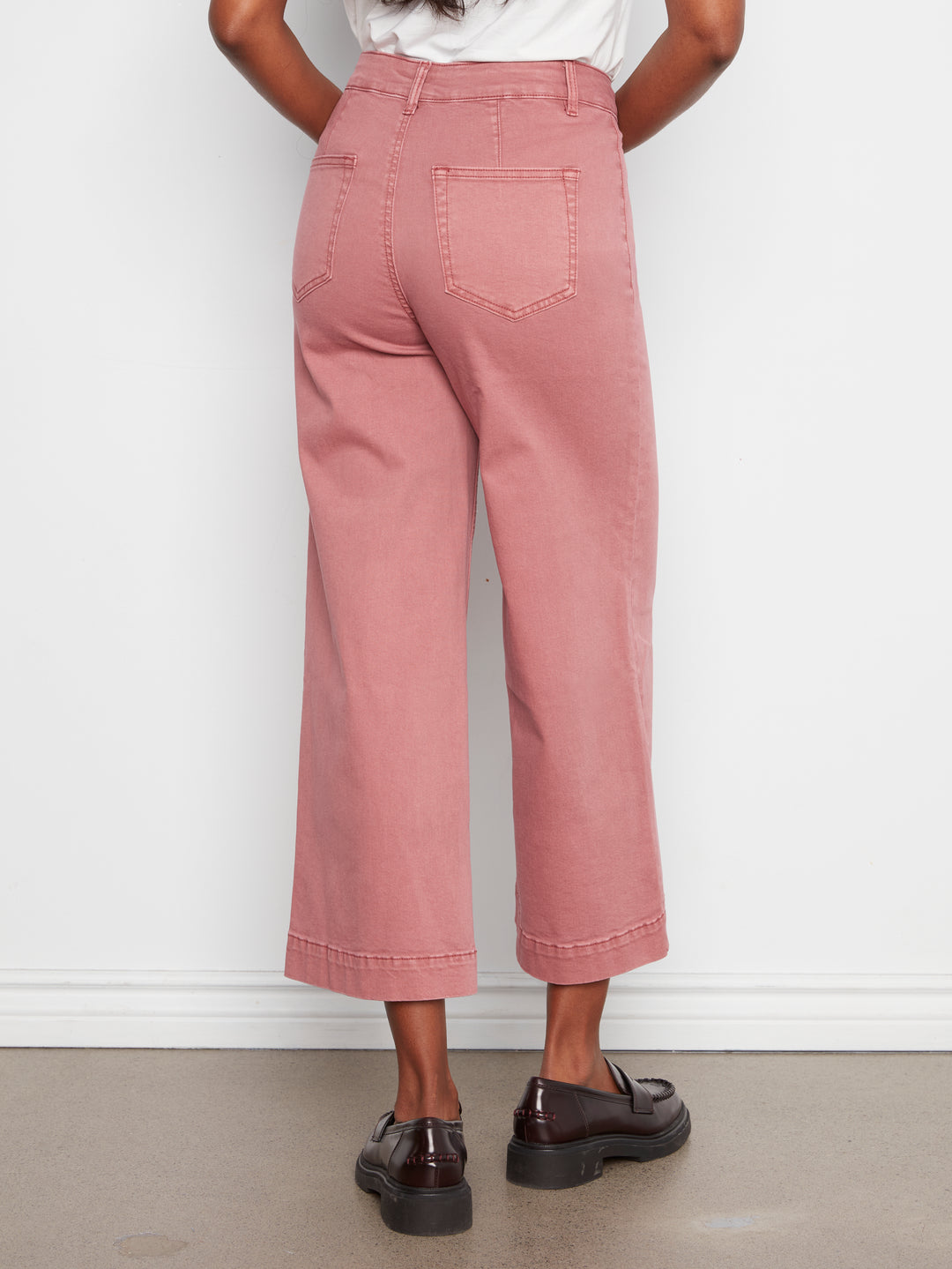 A person shown from the back wearing dusty rose cropped wide-leg jeans with rolled hems, paired with a white top and dark brown leather loafers, standing against a white wall.