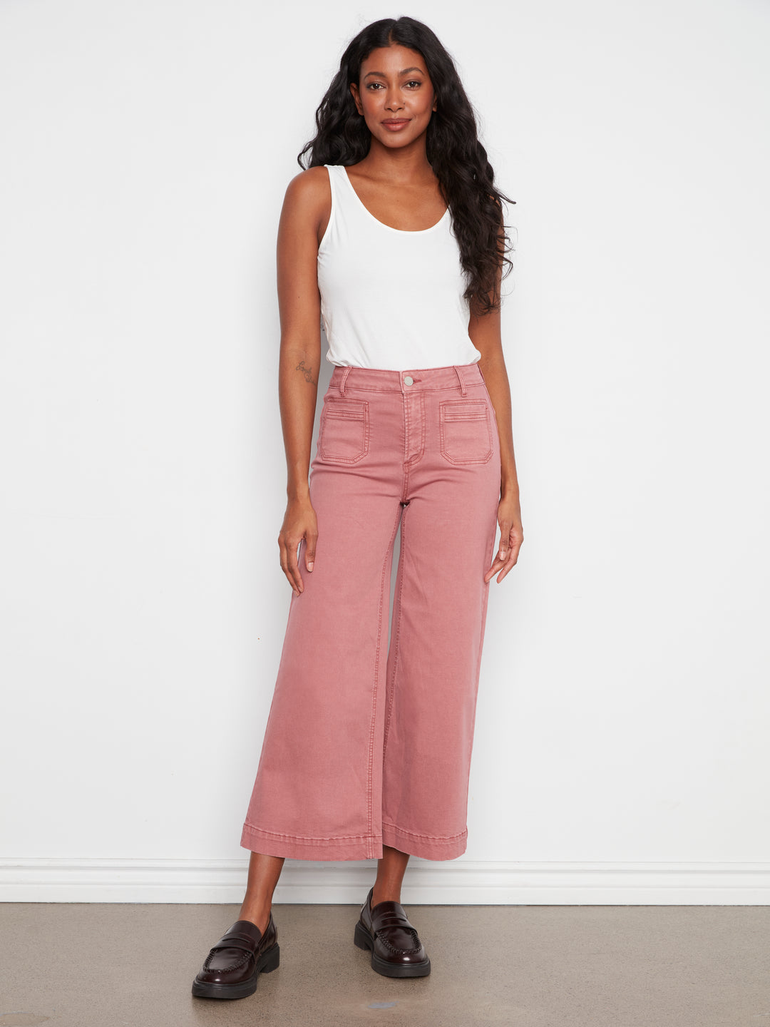 A woman modeling dusty rose wide-leg cropped jeans with a white tank top and black loafers, standing against a plain white wall.