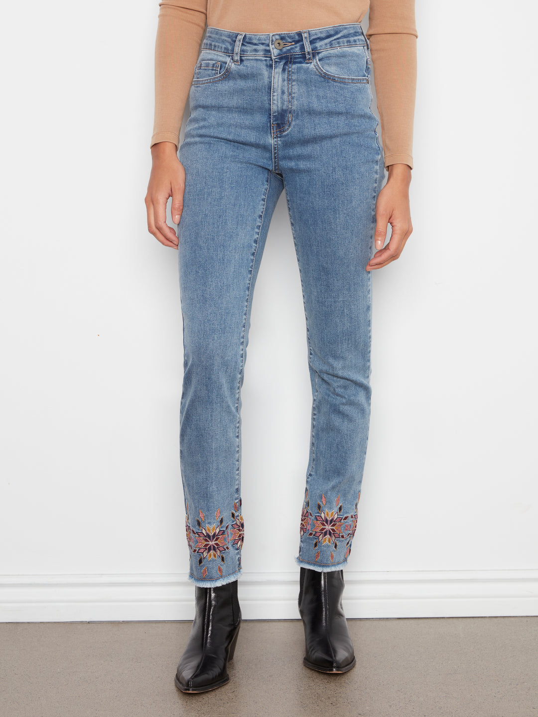 A person wearing light blue high-waisted jeans with colorful floral embroidery near the hems, paired with a tan long-sleeve top and black ankle boots, standing against a white wall.