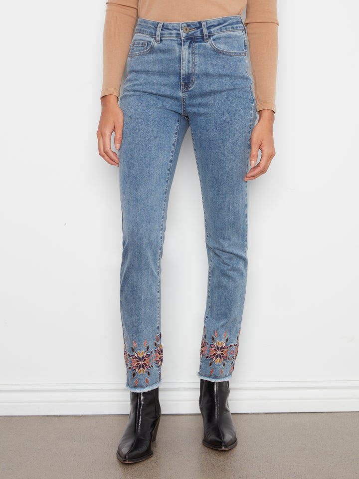A person wearing light blue high-waisted jeans with colorful floral embroidery near the hems, paired with a tan long-sleeve top and black ankle boots, standing against a white wall.