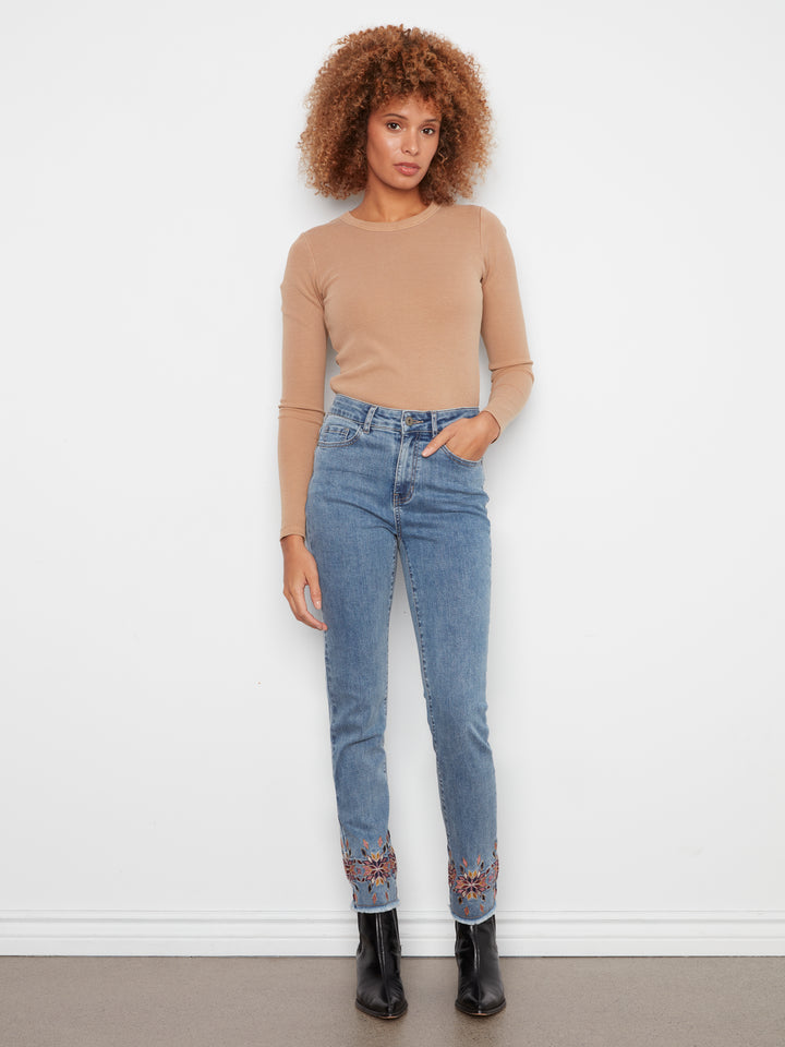 A woman stands against a white wall wearing a beige long-sleeve top, light blue jeans with decorative embroidery at the cuffs, and black ankle boots.