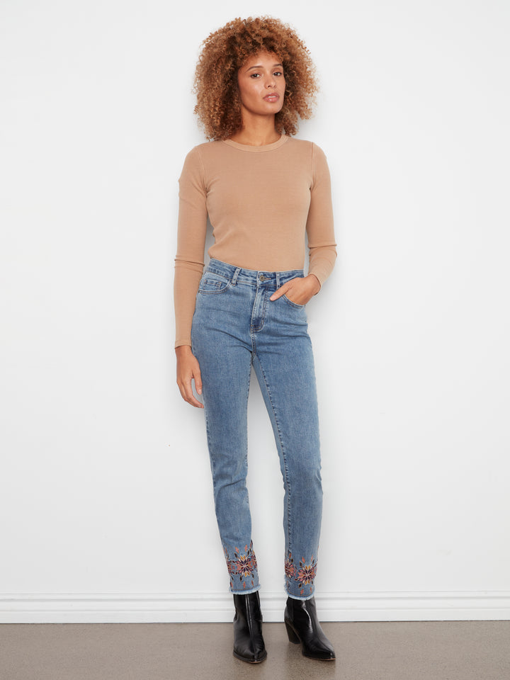 A woman modeling a beige long-sleeve knit top and light blue high-waisted skinny jeans with lace-up ankle details, paired with black ankle boots, standing against a white wall.