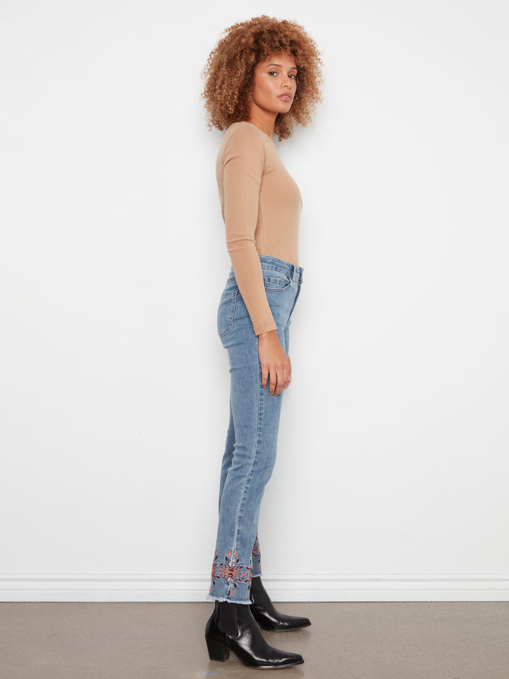 A woman stands in profile wearing a beige long-sleeve top, light blue jeans with floral embroidery near the hem, and black ankle boots against a plain white wall.