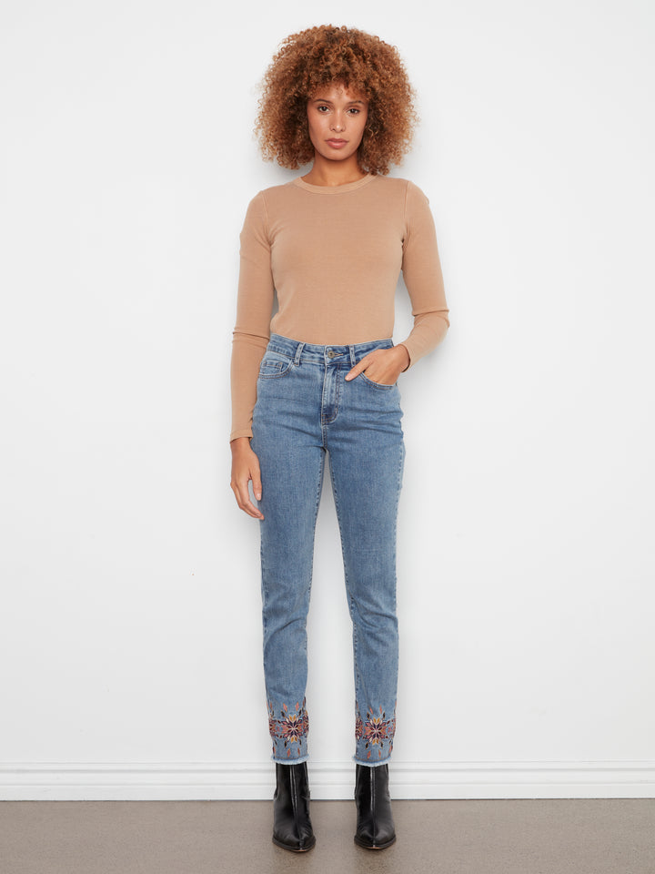 A woman stands against a white wall wearing a beige long-sleeve top, light blue jeans with embroidered detailing at the cuffs, and black ankle boots.
