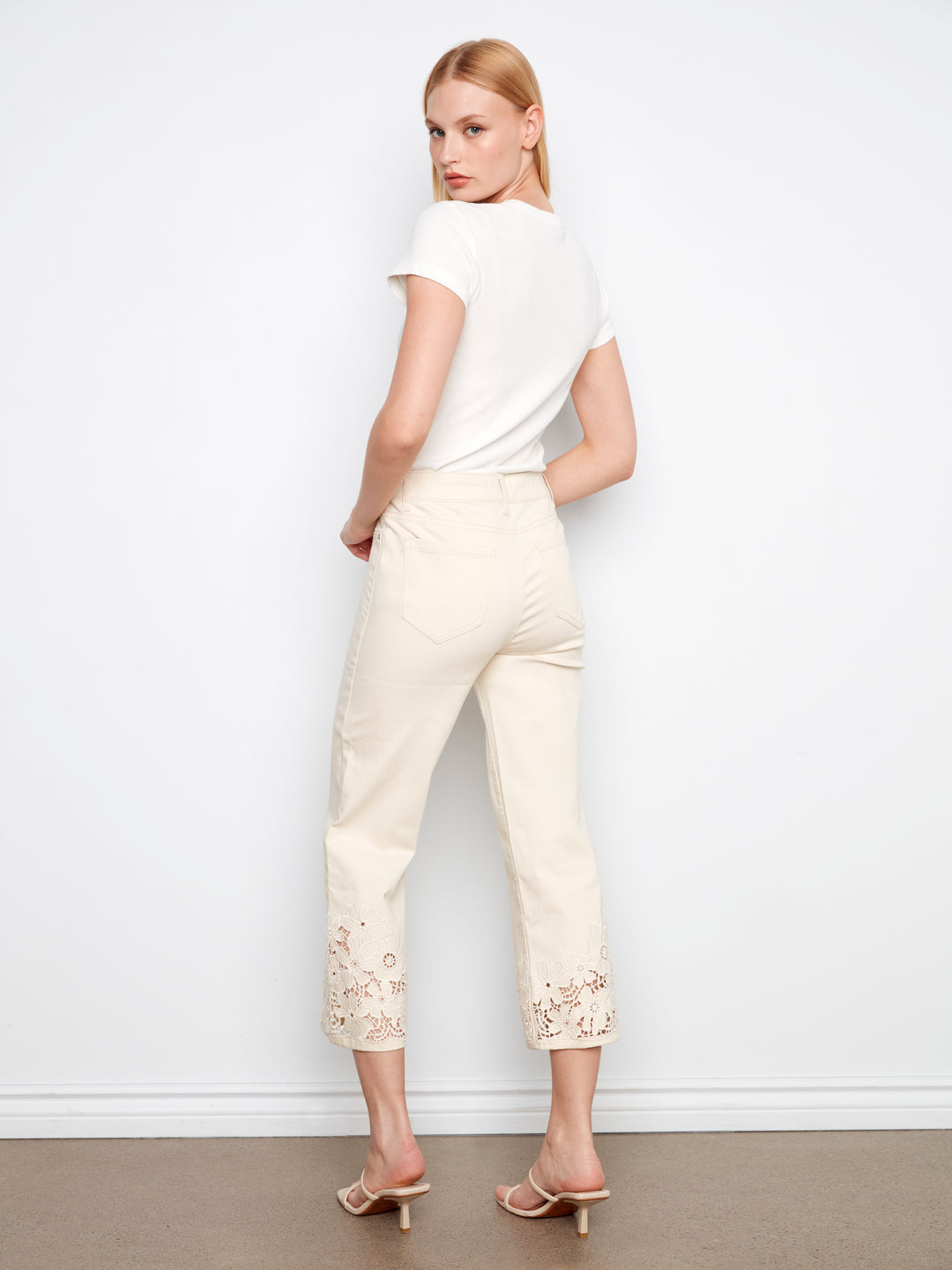 Woman wearing a white top and cream pants with lace details against a plain background