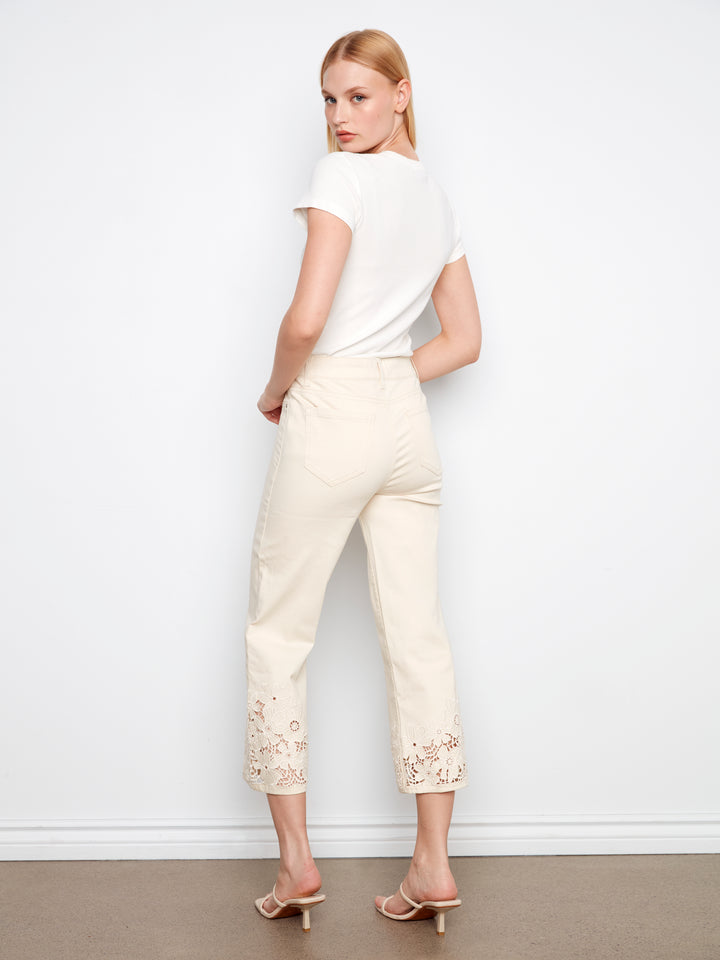 Woman wearing a white top and cream pants with lace details against a plain background