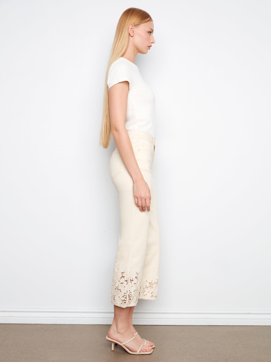 Woman wearing a white top and cream pants with floral details against a plain background