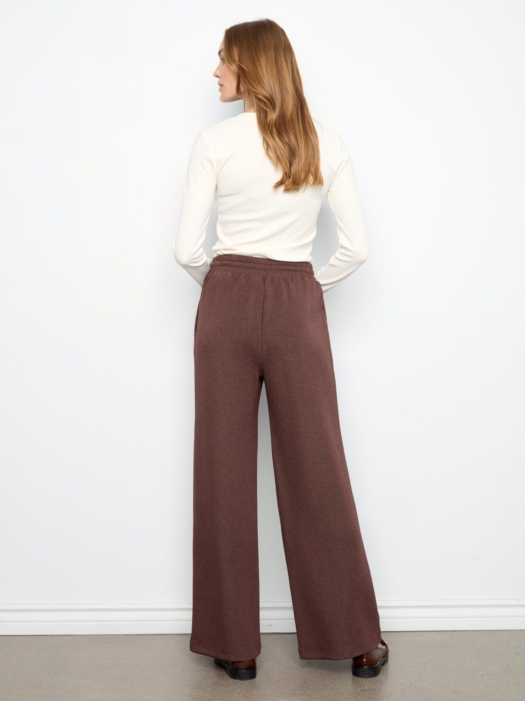 A woman shown from the back wearing a white long-sleeve top and high-waisted, wide-leg brown pants against a plain white wall.