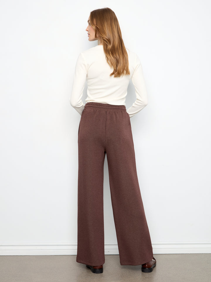 A woman shown from the back wearing a white long-sleeve top and high-waisted, wide-leg brown pants against a plain white wall.