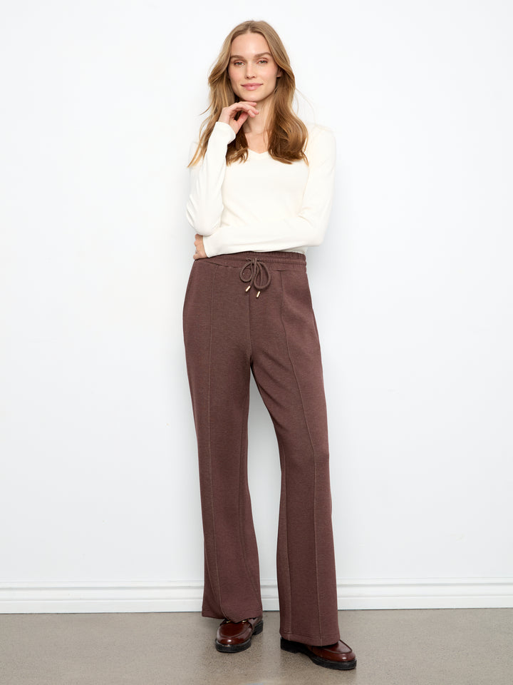 A woman modeling a pair of brown wide-leg drawstring pants with a cream long-sleeve top and dark brown leather shoes, standing against a white wall.