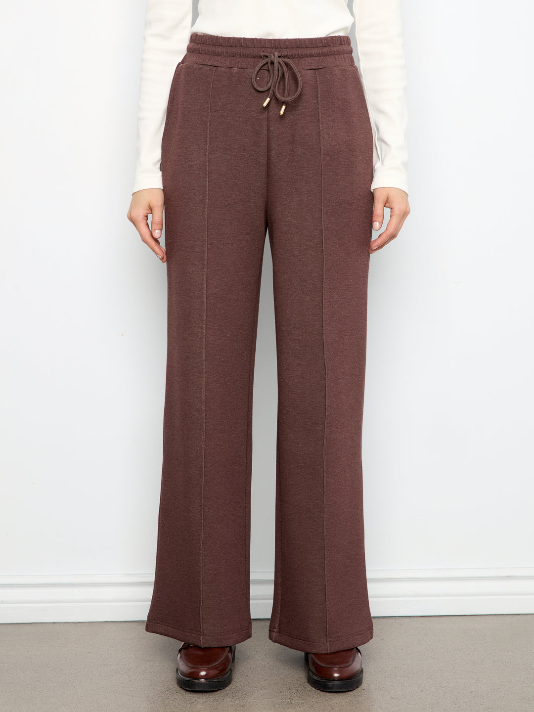 A person wearing brown wide-leg drawstring pants with a ribbed knit top and brown shoes, standing against a white wall.