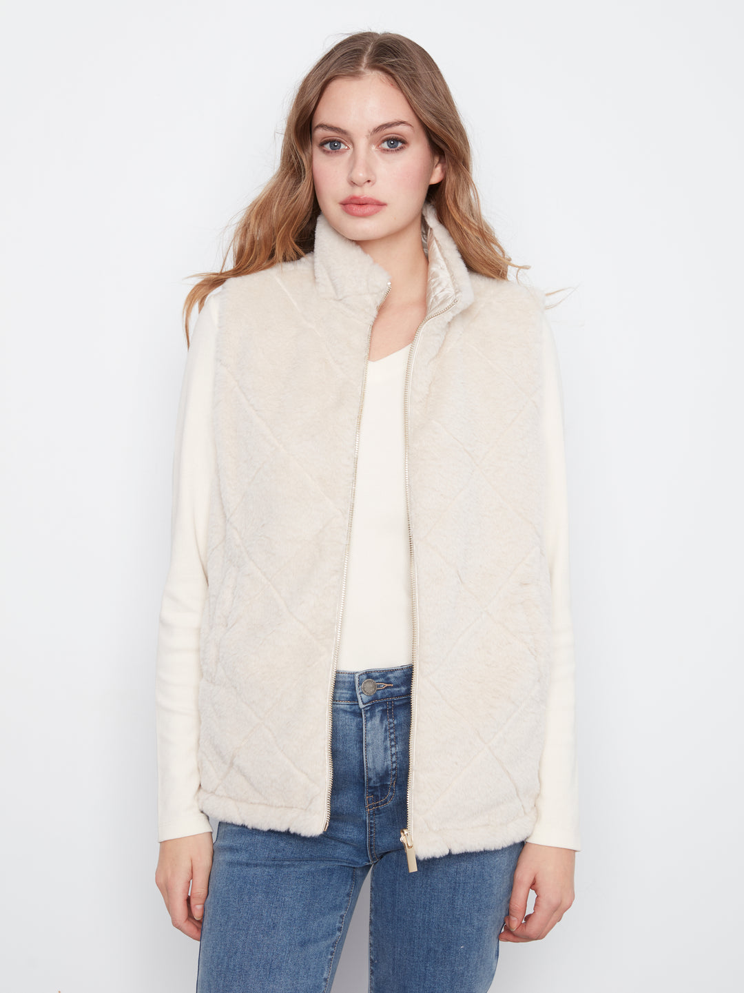 A woman modeling a cream-colored, diamond-quilted faux-fur vest over a cream top and blue jeans, photographed against a plain white studio background.