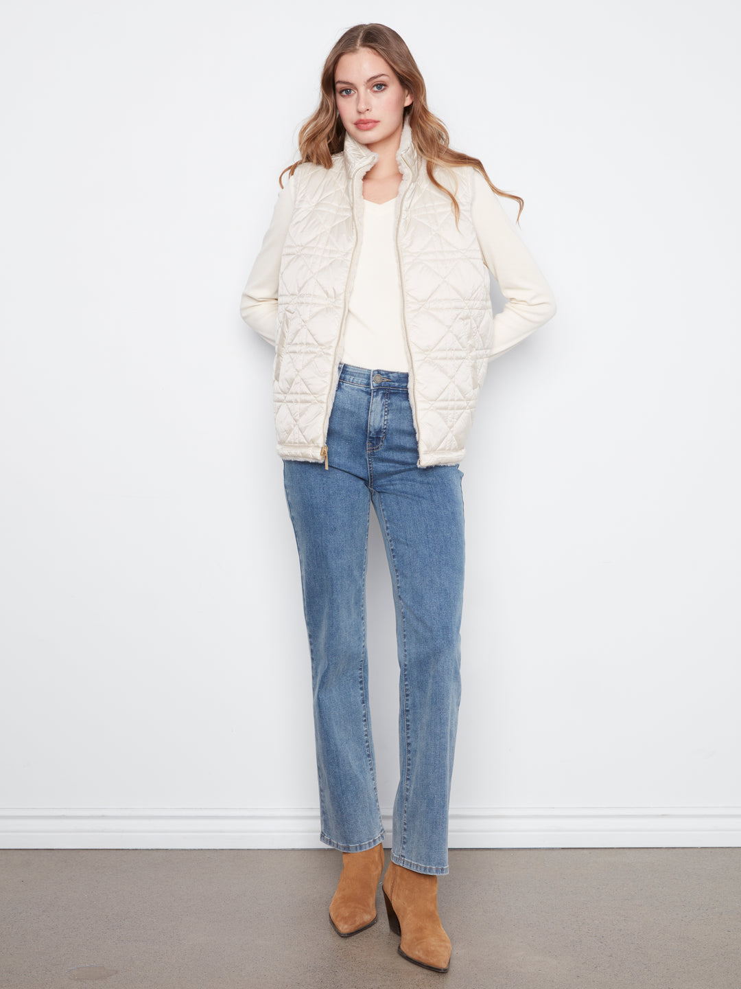 A woman modeling a cream quilted zip-up puffer jacket over a cream top, paired with blue jeans and tan ankle boots, standing against a white wall.
