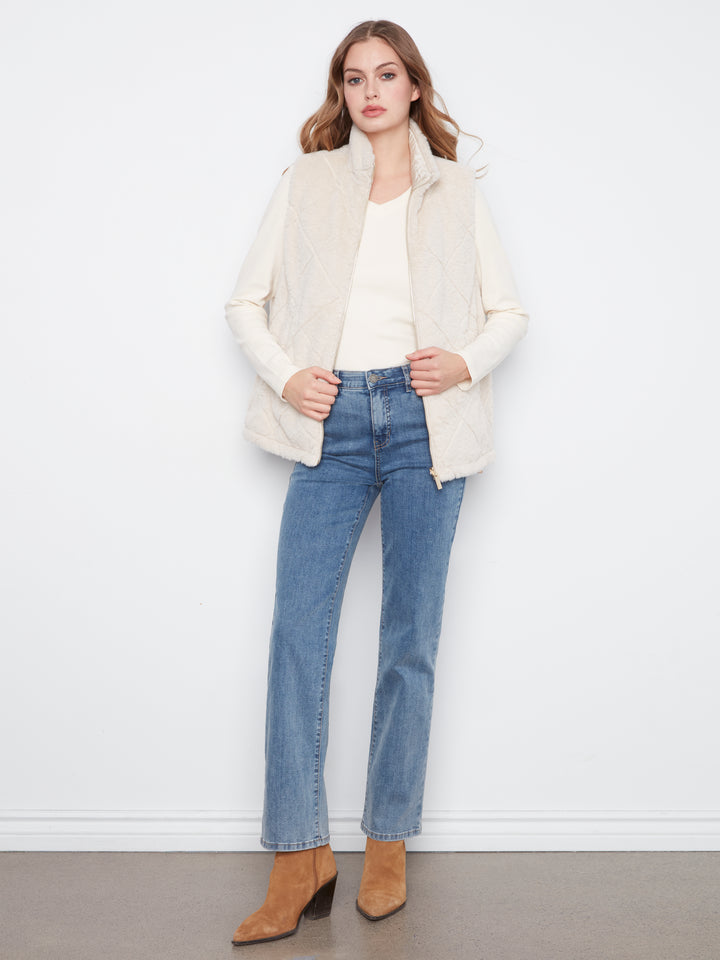 A woman stands against a white wall wearing a cream sleeveless quilted vest over a cream top, blue straight-leg jeans, and tan ankle boots.