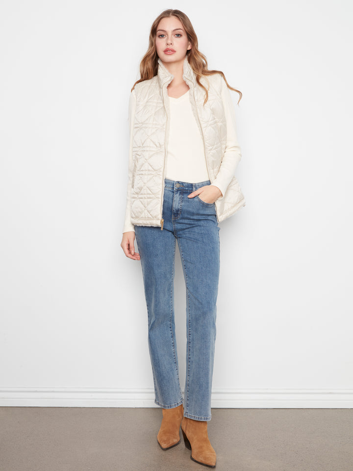 A woman stands against a white wall wearing a cream quilted zip-front vest over a cream top, blue jeans, and tan ankle boots.