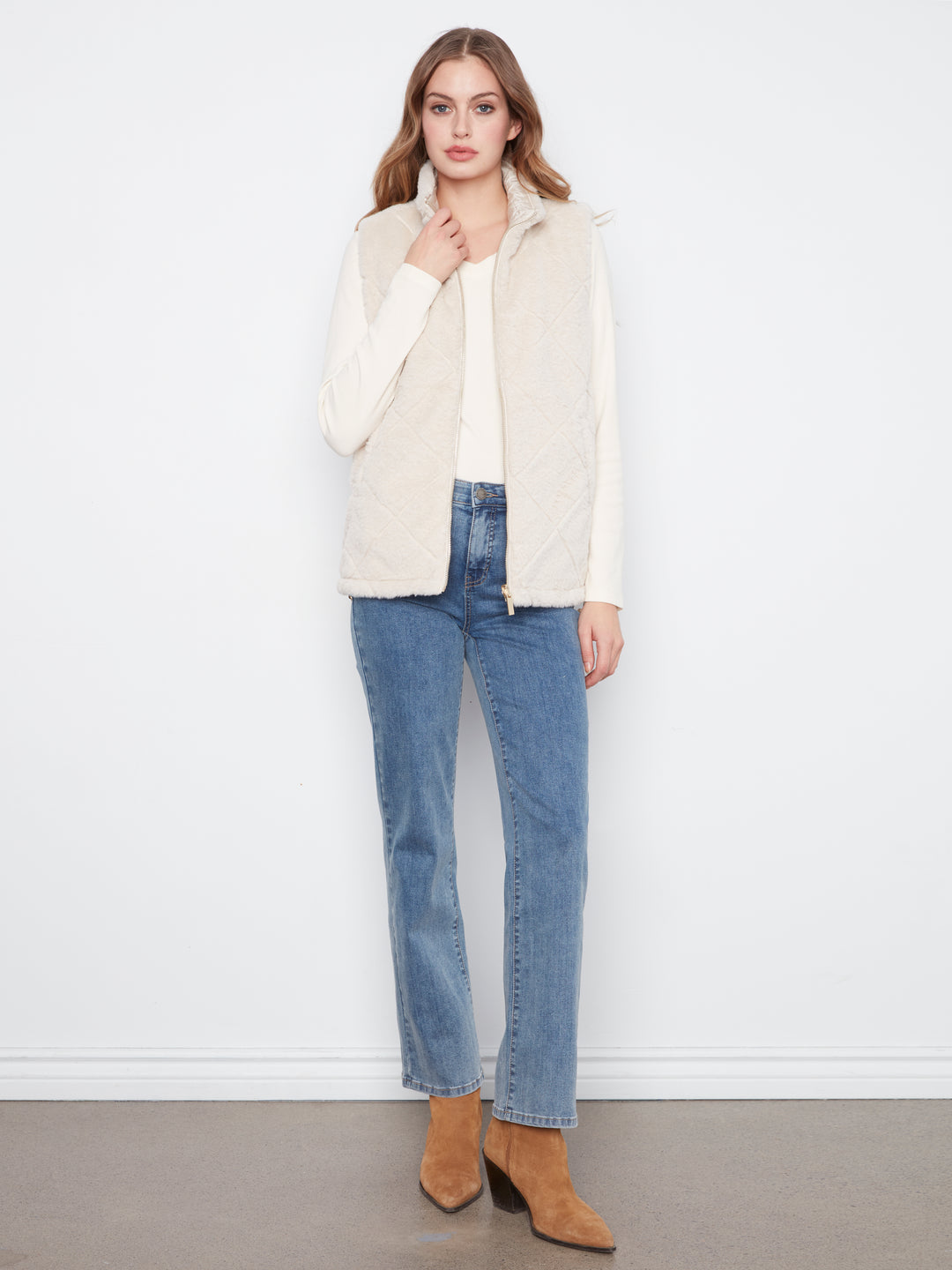 A woman stands against a white wall wearing a cream quilted sleeveless vest over a white top, blue jeans, and tan ankle boots.