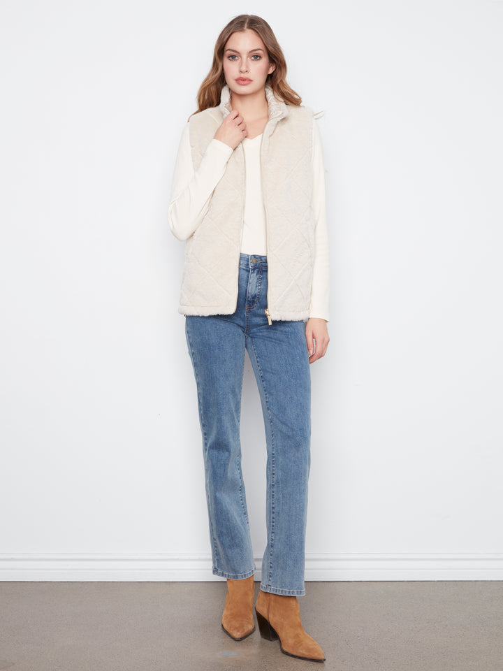 A woman stands against a white wall wearing a cream quilted sleeveless vest over a white top, blue jeans, and tan ankle boots.