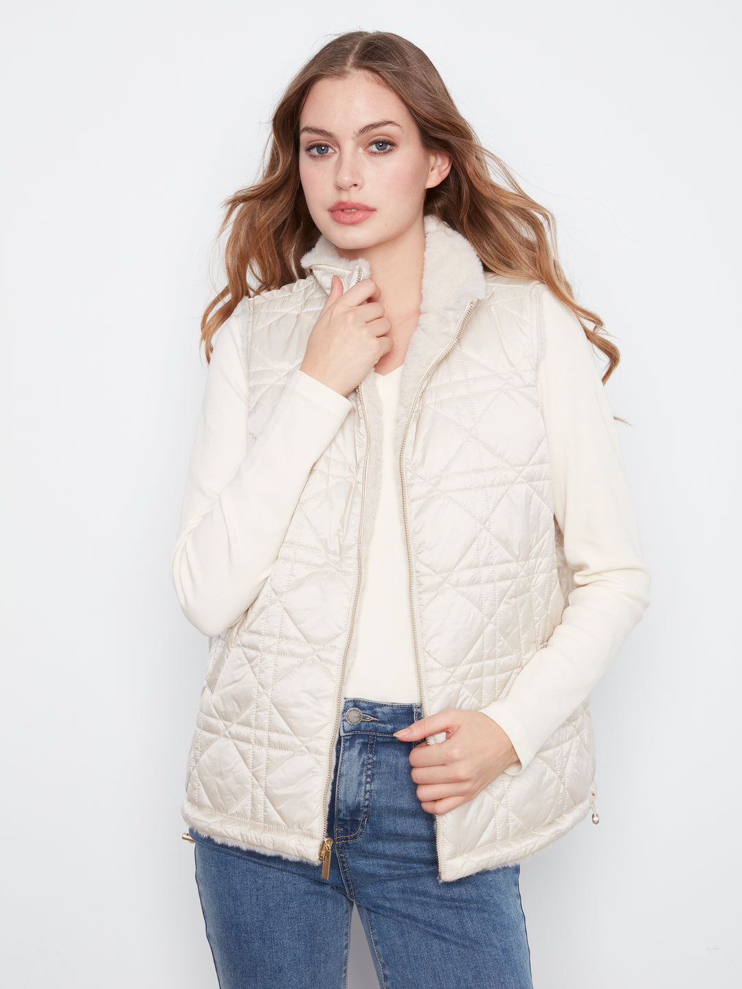 A woman modeling a cream-colored quilted puffer vest with a fuzzy collar, worn over a matching cream top and blue jeans, against a plain white studio background.