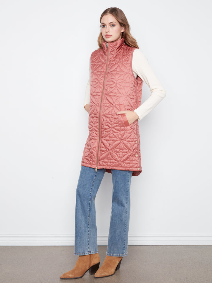 A woman modeling a dusty rose pink quilted sleeveless puffer vest over a white long-sleeve top, blue jeans, and tan ankle boots, standing against a plain white wall.