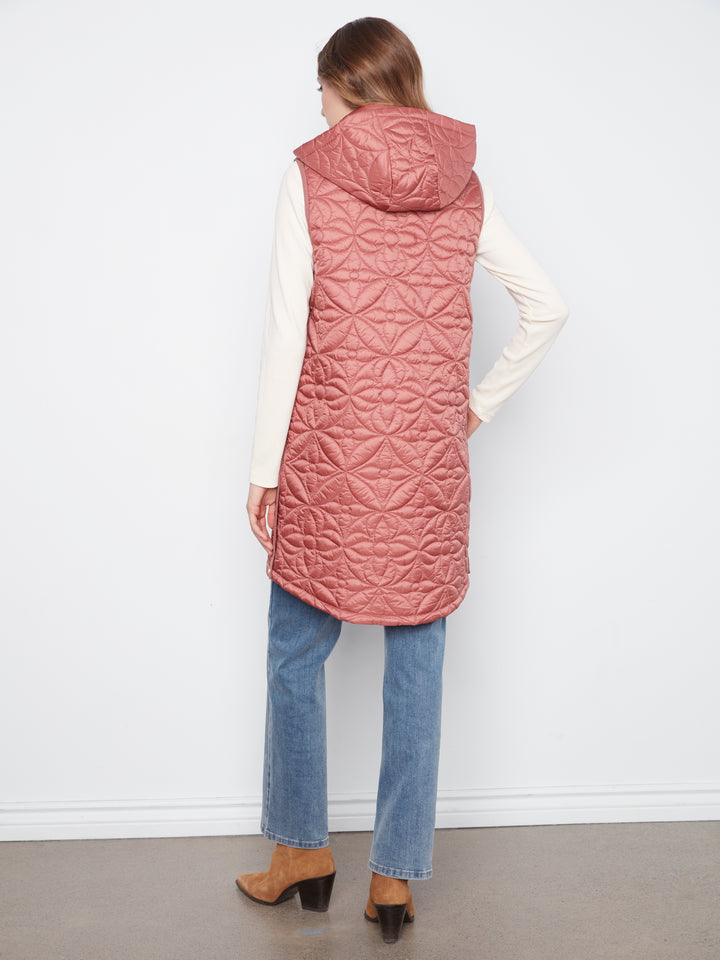 A woman shown from the back wearing a dusty rose pink quilted sleeveless hooded puffer vest over a cream long-sleeve top, blue jeans, and brown ankle boots, standing against a white wall.